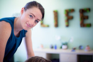 A chiropractor, a white woman wearing a blue shirt, leans over a patient to administer an adjustment. The words "LIFE" appear in the out-of-focus background.