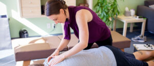  A white woman, with brown hair, is shown giving a chiropractic adjustment to a man who is lying face-down on a chiropractic table.